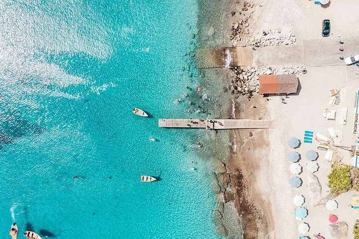 Vista aérea de una playa con un muelle de madera, aguas claras y barcos.